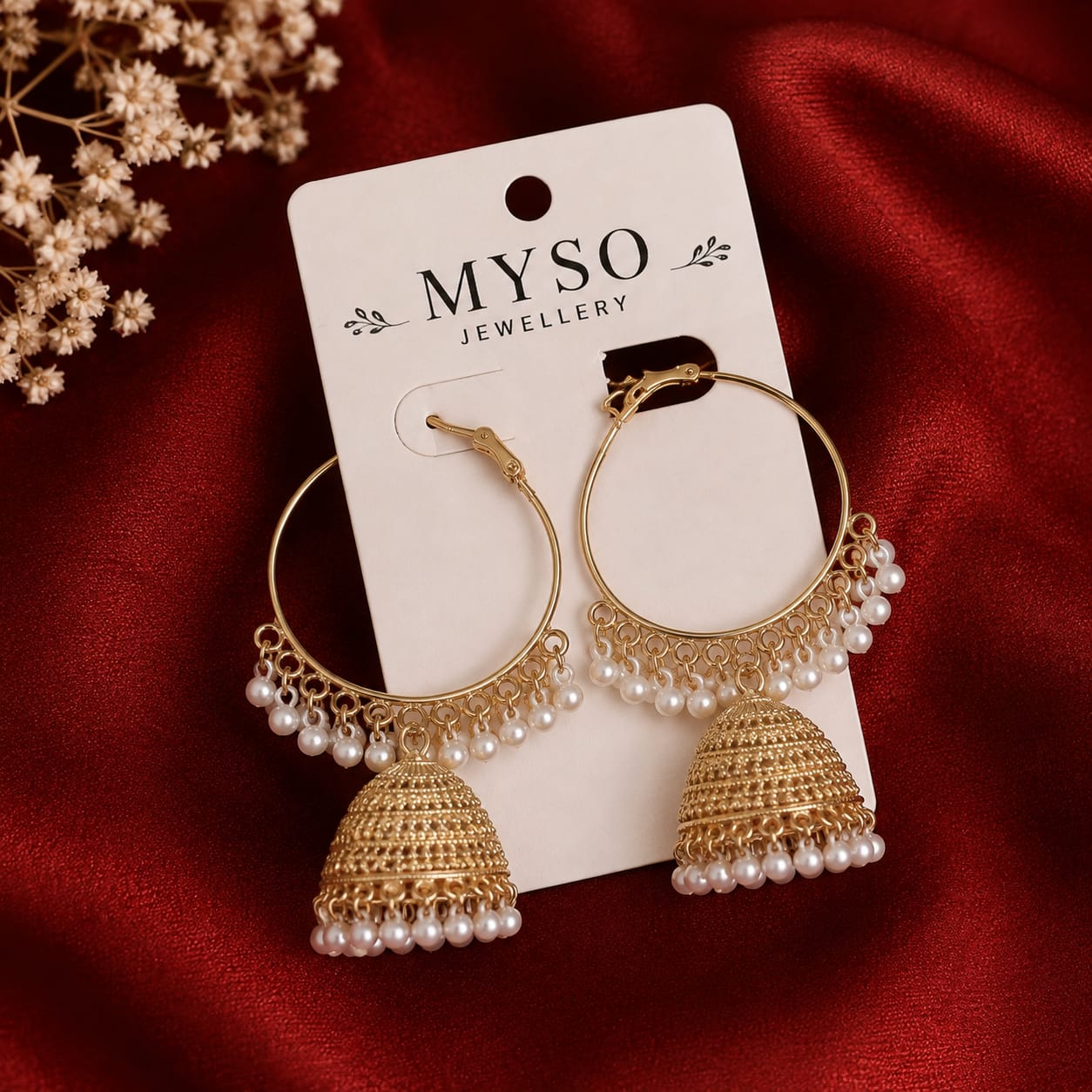 A pair of MYSO JEWELLERY gold-tone hoop earrings with dangling bell-shaped jhumka-style charms and rows of white pearls, displayed on a white branded card over a rich red satin fabric with dried flowers in the corner.